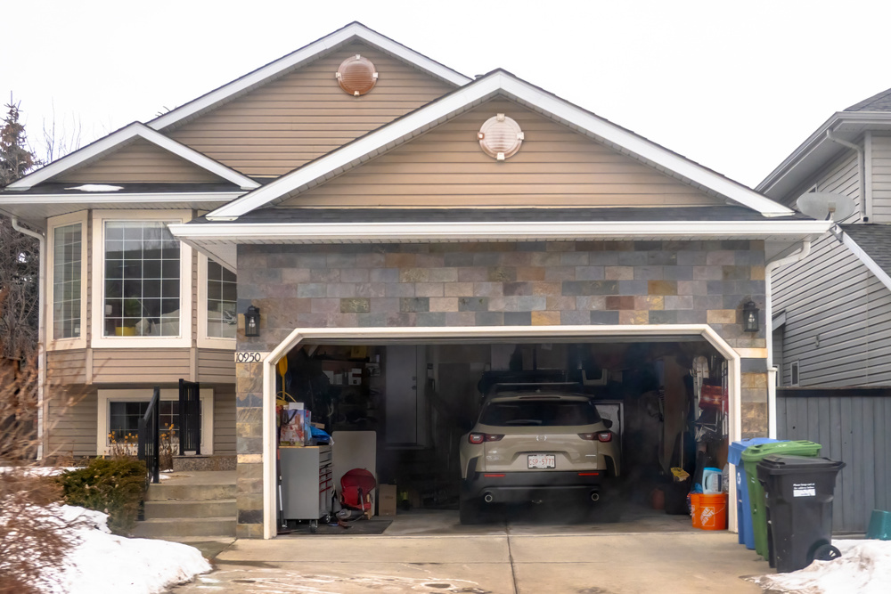 Calgary, Alberta, Canada. January 15, 2026. A suburban detached house in winter with an open garage showing a parked SUV, household storage, and recycling bins